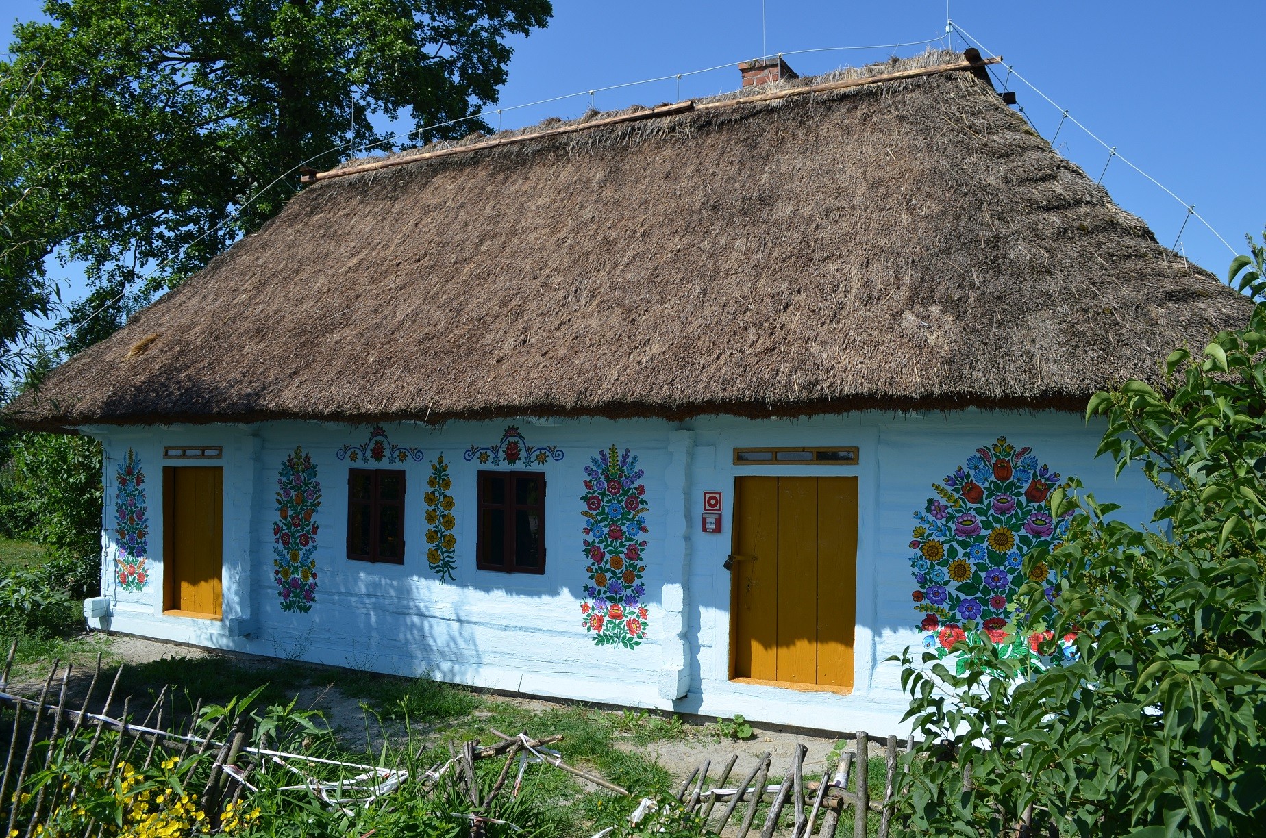 Drewniana chałupa pomalowana na biało kryta strzechą. Na ścianach domu są wielobarwne kompozycje kwiatowe.